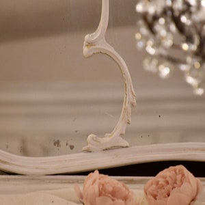 May include: A white ornate mirror with a decorative, carved frame. The mirror is reflecting a chandelier with crystal accents.