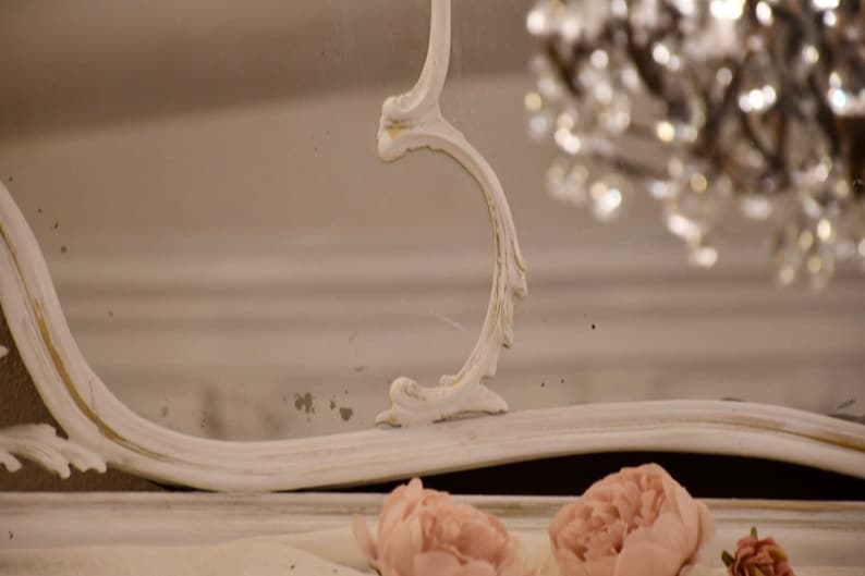 May include: A white ornate mirror with a decorative, carved frame. The mirror is reflecting a chandelier with crystal accents.