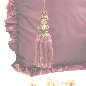 May include: A purple tassel with a gold metal top. The tassel is hanging from a purple fabric with ruffles.