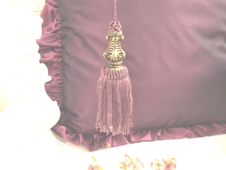 May include: A purple tassel with a gold metal top. The tassel is hanging from a purple fabric with ruffles.