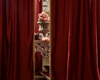 May include: Deep burgundy velvet curtains with gold trim. A glimpse of a decorative interior is visible. A gold chandelier hangs in the mirror's reflection. A white ornate table holds a vase of pink flowers, creating an elegant ambiance.