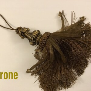 May include: A brown tassel with a gold-colored metal top. The tassel is made of a soft, fluffy material and has a long, thin cord.