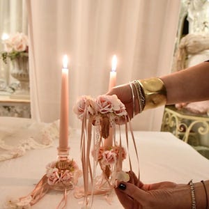 May include: Two lit pink candles in decorative glass holders, embellished with pink flowers and ribbons. A hand holds a floral arrangement with ribbons, matching the candle decor. The scene suggests a romantic theme.