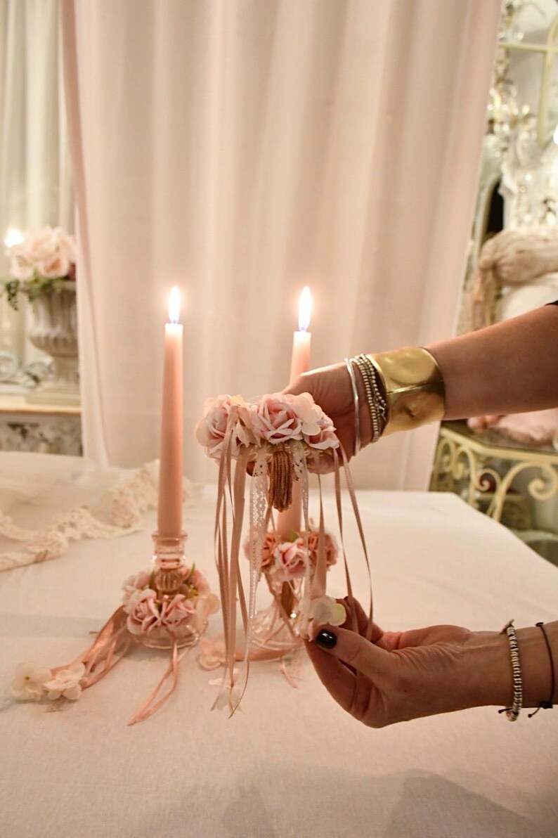 May include: Two lit pink candles in decorative glass holders, embellished with pink flowers and ribbons. A hand holds a floral arrangement with ribbons, matching the candle decor. The scene suggests a romantic theme.