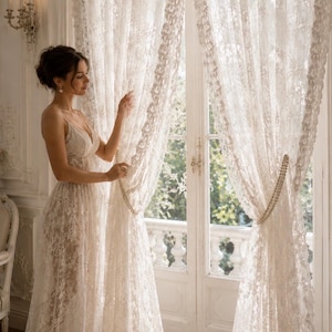 May include: Floor-length white lace curtains featuring an intricate floral pattern and sheer texture. The curtains are gathered at the sides with beaded tiebacks, allowing light to filter through the delicate fabric into the room.