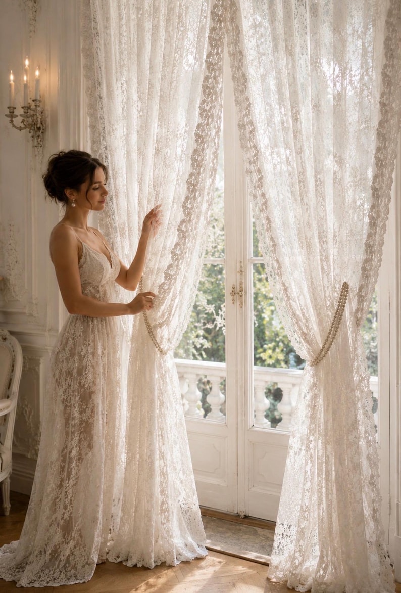 May include: Floor-length white lace curtains featuring an intricate floral pattern and sheer texture. The curtains are gathered at the sides with beaded tiebacks, allowing light to filter through the delicate fabric into the room.