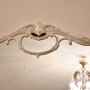 May include: A white ornate mirror with a decorative, carved frame. The mirror is reflecting a chandelier with crystal accents.