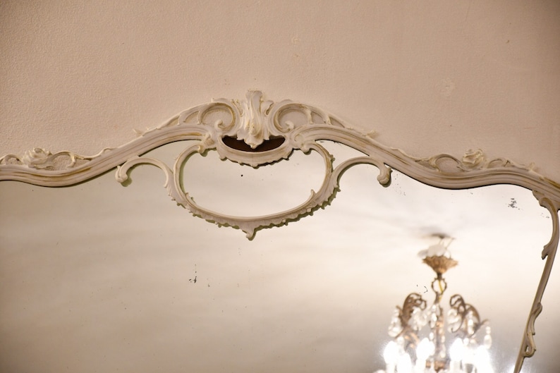 May include: A white ornate mirror with a decorative, carved frame. The mirror is reflecting a chandelier with crystal accents.