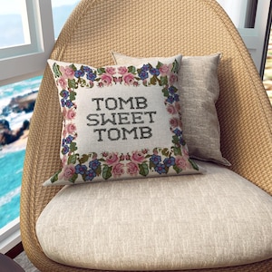 May include: A decorative pillow with the text "TOMB SWEET TOMB" in black cross-stitch. The pillow is surrounded by a floral border of pink roses, blue flowers, and green leaves. A second, solid-colored pillow is behind it.