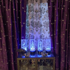May include: Four glass light-up figurines of characters from the Haunted Mansion attraction at Disney World. The figurines are on a shelf with other Halloween decorations. The figurines are lit up with blue light. The shelf is in front of a window with purple curtains and a sheer curtain with a black and white pattern.