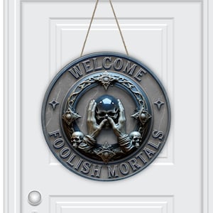 May include: A round, gray metal welcome sign with the words "WELCOME FOOLISH MORTALS" in a gothic font. The sign features a skull held by hands, surrounded by skulls and decorative elements, hanging from a brown string.