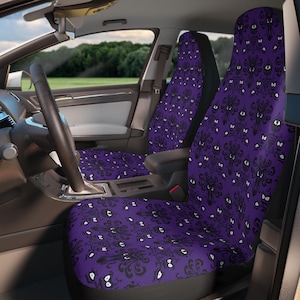 May include: Purple car seat covers with a black and white haunted house pattern. The covers feature a repeating design of stylized haunted houses and white, glowing eyes. The interior of a car is visible, with the steering wheel and dashboard in view.