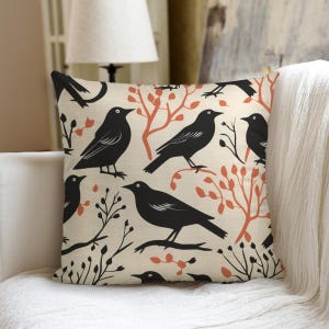 Puede incluir: Cojín cuadrado con un diseño de cuervos negros y ramas sobre un fondo beige. El diseño incluye ramas de color naranja rojizo y follaje negro, creando un tema inspirado en la naturaleza. El cojín probablemente está hecho de una tela suave.