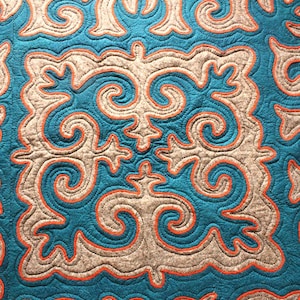 May include: Close-up of a textile with a repeating pattern. The design features teal, grey, and orange colours. The teal sections are outlined with orange stitching, and the grey sections have a similar orange outline.