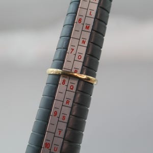 May include: A gold ring is placed on a ring sizer. The ring is a simple band design. The ring sizer is dark gray with white markings for ring sizes. The ring appears to be a size 8.