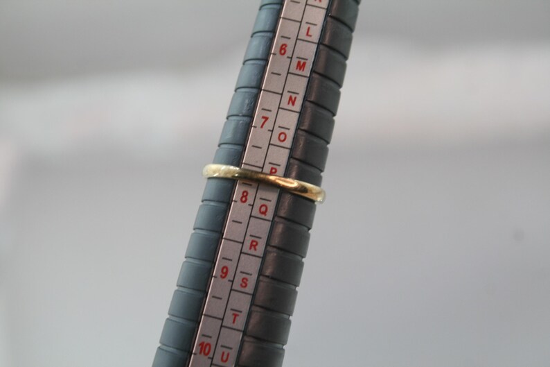 May include: A gold ring is placed on a ring sizer. The ring is a simple band design. The ring sizer is dark gray with white markings for ring sizes. The ring appears to be a size 8.