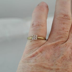 May include: A gold-toned ring with a square cluster of small, clear stones. The ring is set on a finger, showcasing the delicate design and the sparkle of the gemstones. The band is thin and simple.