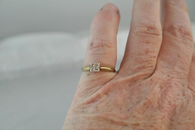 May include: A gold-toned ring with a square cluster of small, clear stones. The ring is set on a finger, showcasing the delicate design and the sparkle of the gemstones. The band is thin and simple.