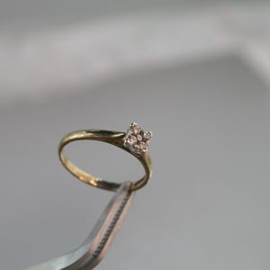 May include: A gold ring with a square cluster of small, clear gemstones. The ring is held by a pair of silver tweezers against a blurred background. The ring's band is thin and yellow.