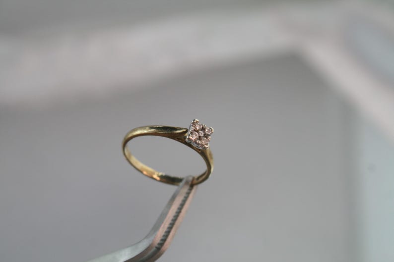 May include: A gold ring with a square cluster of small, clear gemstones. The ring is held by a pair of silver tweezers against a blurred background. The ring's band is thin and yellow.
