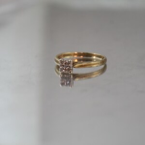 May include: A gold ring with a square cluster of small, clear gemstones. The ring is set on a reflective surface, creating a mirrored effect. The band is thin and appears to be made of gold.