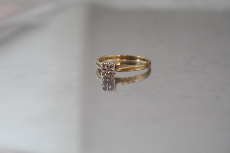 May include: A gold ring with a square cluster of small, clear gemstones. The ring is set on a reflective surface, creating a mirrored effect. The band is thin and appears to be made of gold.
