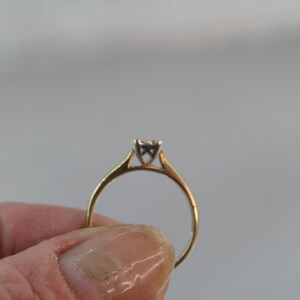 May include: A gold ring with a small, square-cut diamond. The ring has a simple, classic design with a thin band and a four-prong setting. The diamond appears to be approximately 0.15 carats.