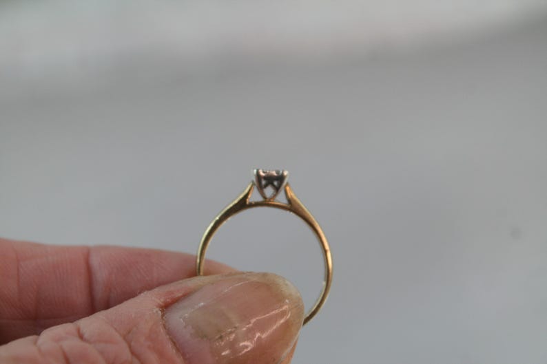 May include: A gold ring with a small, square-cut diamond. The ring has a simple, classic design with a thin band and a four-prong setting. The diamond appears to be approximately 0.15 carats.