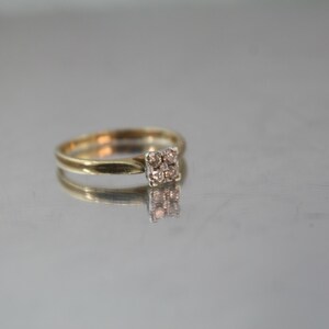 May include: A gold ring with a square cluster of small, light-colored stones. The ring is set on a reflective surface, creating a mirrored effect. The band is thin and simple, highlighting the central stone arrangement.