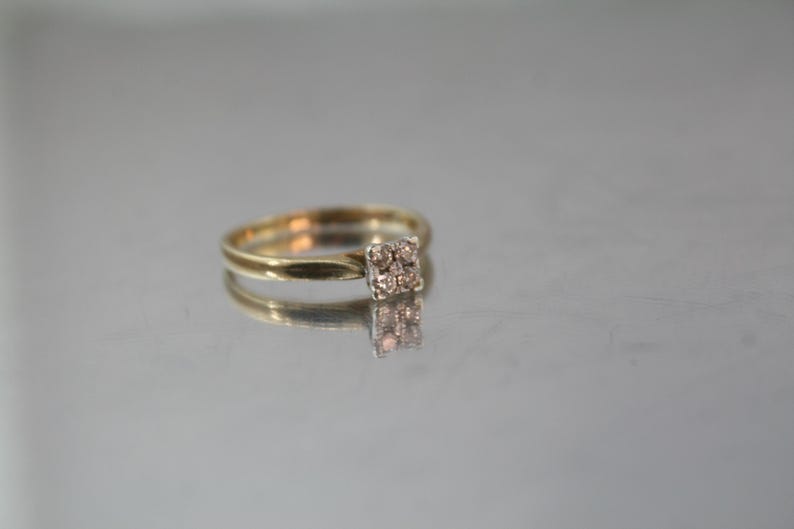 May include: A gold ring with a square cluster of small, light-colored stones. The ring is set on a reflective surface, creating a mirrored effect. The band is thin and simple, highlighting the central stone arrangement.