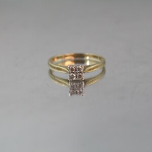 May include: A gold ring with a square cluster of small, light-colored stones at the center. The ring is set against a reflective surface, creating a mirrored effect. The band is a simple, elegant design.