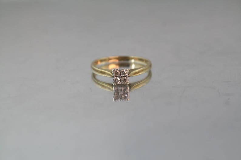 May include: A gold ring with a square cluster of small, light-colored stones at the center. The ring is set against a reflective surface, creating a mirrored effect. The band is a simple, elegant design.