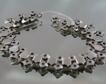 May include: Silver bracelet with a delicate, intricate design. The bracelet is made up of small, interconnected links that form a repeating pattern. The bracelet is shown on a reflective surface.