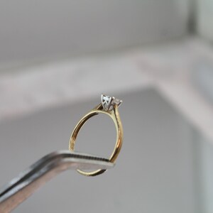 May include: A gold engagement ring with a single, clear diamond held by a pair of silver tweezers. The ring has a classic design with a slender band and a four-prong setting. The diamond is the focal point.
