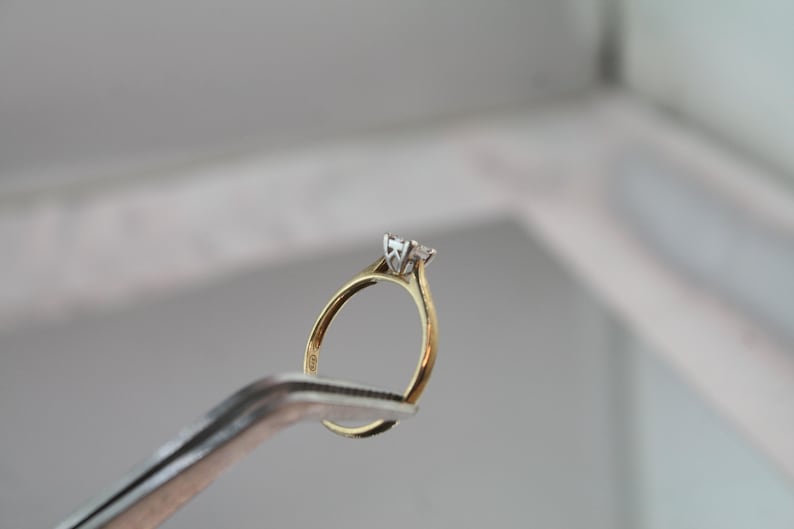 May include: A gold engagement ring with a single, clear diamond held by a pair of silver tweezers. The ring has a classic design with a slender band and a four-prong setting. The diamond is the focal point.