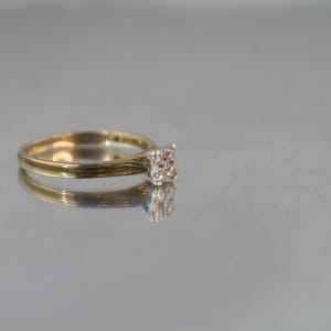 May include: A gold ring with a square cluster of small, clear stones. The ring is set on a reflective surface, creating a mirrored effect. The band is thin and polished, and the stones are set close together.