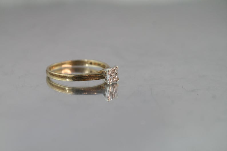 May include: A gold ring with a square cluster of small, clear stones. The ring is set on a reflective surface, creating a mirrored effect. The band is thin and polished, and the stones are set close together.