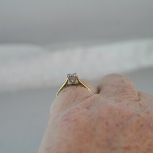 May include: A gold ring with a square-cut diamond set in a four-prong setting. The ring is held up against a light background, showcasing the diamond's sparkle. The ring's band is a simple, classic design.