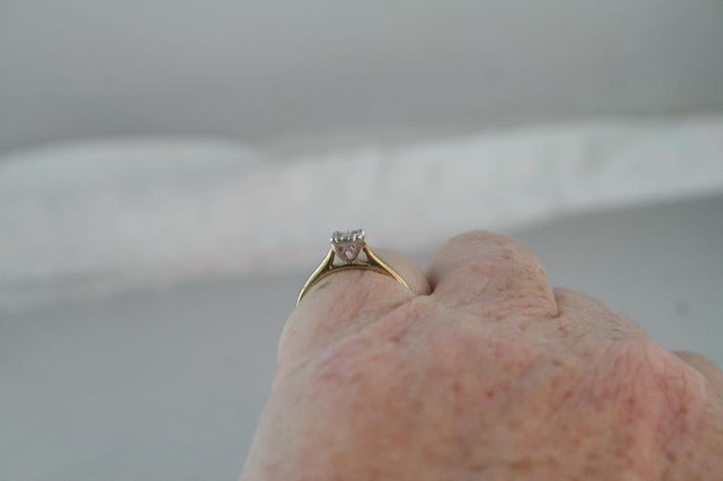 May include: A gold ring with a square-cut diamond set in a four-prong setting. The ring is held up against a light background, showcasing the diamond's sparkle. The ring's band is a simple, classic design.