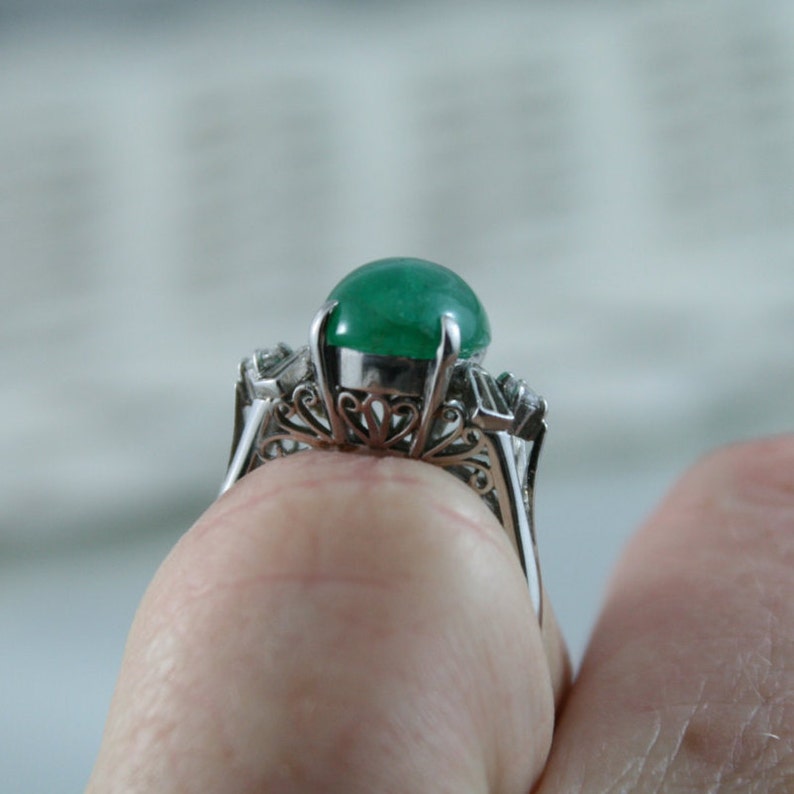 May include: A silver ring with a large, round, green emerald gemstone set in a filigree design. The ring is set with smaller diamonds on either side of the emerald.