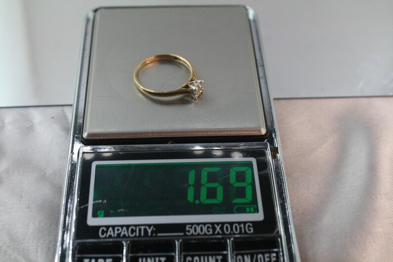 May include: A gold ring with a diamond-shaped stone is displayed on a digital scale. The scale shows a weight of 1.69, with a capacity of 500g x 0.01g. The ring is centered on the scale's silver surface.