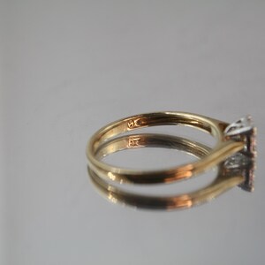 May include: A gold ring with a square-shaped diamond cluster. The ring is set on a reflective surface, showcasing its design. The band is a simple, classic style.