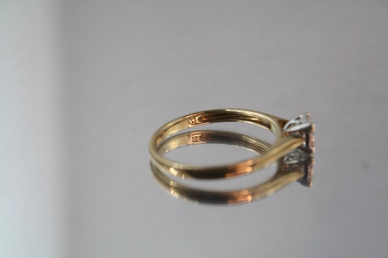 May include: A gold ring with a square-shaped diamond cluster. The ring is set on a reflective surface, showcasing its design. The band is a simple, classic style.