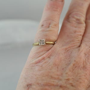 May include: A gold ring with a square cluster of small diamonds. The ring is on a finger, with the hand in the foreground. The background is blurred and light-colored.