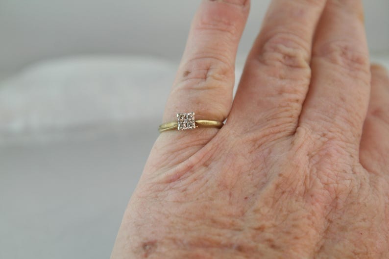 May include: A gold ring with a square cluster of small diamonds. The ring is on a finger, with the hand in the foreground. The background is blurred and light-colored.