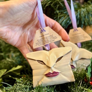Può includere: Ornamento in legno chiaro a forma di busta con un volto, occhi marroni e labbra rosse. È appeso a un nastro viola e a un'etichetta con testo. Sfondo con vegetazione verde.