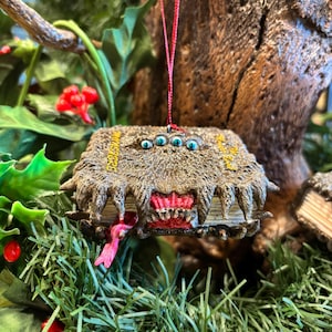 May include: A detailed, fantasy-themed Christmas ornament shaped like a monster book. The book is brown with a textured, furry appearance, featuring a red mouth with teeth and three blue eyes. A pink tongue extends from the mouth.