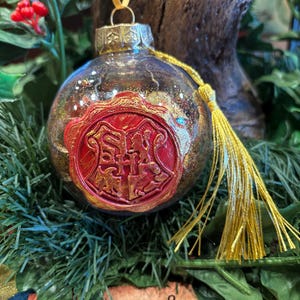 May include: A clear glass Christmas ornament with gold glitter and a red and gold emblem. The ornament has a gold tassel and ribbon, and is surrounded by greenery and berries. The emblem features a crest with a shield and two animals.