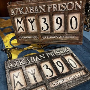 Puede incluir: Dos letreros de madera con cadenas metálicas colgando de la parte superior. Los letreros son de color marrón oscuro y tienen un aspecto desgastado. Los letreros dicen "Azkaban Prison" y tienen una pantalla de números con el número "390". Los letreros tienen una serie de símbolos y números debajo de la pantalla de números.