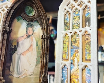 May include: Two decorative ornaments shaped like stained glass windows. One ornament features a woman in a white gown with a green wreath on her head. The other ornament features a stained glass design with a woman in a blue gown.